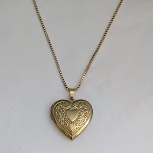May include: A gold-coloured heart-shaped locket necklace. The locket features an ornate floral design and a small heart in the centre. The necklace chain is a delicate box chain style.