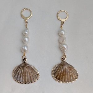 May include: A pair of drop earrings. Each earring features a seashell pendant with a gold-coloured trim, three white, irregular-shaped pearls, and a gold-coloured clasp. The earrings are designed to be worn.