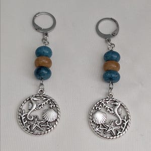 Peut inclure: Boucles d'oreilles pendantes sur le thème de la plage. Chaque boucle d'oreille comporte un charm argenté avec un design d'hippocampe, d'étoile de mer et de coquillage. Les boucles d'oreilles ont également des perles bleues et beiges et des fermoirs à levier argentés.