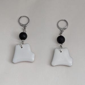 May include: A pair of earrings with white ceramic pendants, each with a red edge, and a black bead. The pendants are suspended from silver-tone lever-back closures. The earrings are handmade and have an irregular shape.