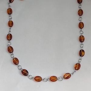 May include: A necklace with oval, amber-coloured beads linked by silver-coloured chain. The translucent beads are strung together, creating a delicate piece of jewellery. The background is white.