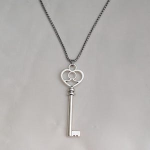 May include: A silver key pendant necklace. The key features a heart-shaped top with a decorative design and a long, straight stem. The chain is a dark silver colour.