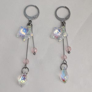 May include: A pair of silver-toned dangle earrings. Each earring features a star-shaped crystal, pink beads, and a teardrop-shaped crystal. The earrings hang from a hoop. The crystals have an iridescent finish.