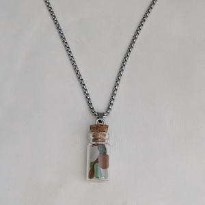 May include: A necklace with a glass bottle pendant containing colourful sea glass. The bottle is sealed with a cork and suspended from a silver-toned chain. The sea glass pieces are in shades of green, blue, and brown.