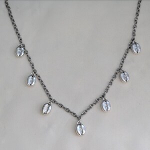 Peut inclure: Un collier argenté avec une chaîne et plusieurs breloques en forme de grains de café. Le collier a un design délicat, avec les breloques en forme de grains de café espacées uniformément le long de la chaîne. Le collier est présenté sur un fond neutre.