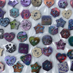 May include: An assortment of colourful, patterned buttons in various shapes, including hearts, stars, and geometric designs. The buttons are made from a variety of materials, including metal, plastic, and wood.