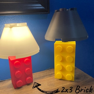 May include: Two lamps with shades, one with a red base and the other with a yellow base. The bases are shaped like large toy building blocks. The lampshades are off-white and gray.