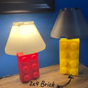 May include: Two lamps with shades, each atop a 2x4 brick base. One lamp has a red base and a cream shade, the other has a yellow base and a dark gray shade. The lamps are on a wooden surface against a blue wall.