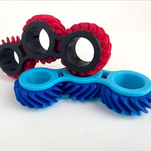 Puede incluir: Dos objetos impresos en 3D. Uno es rojo con anillos negros, el otro es azul. Ambos tienen un diseño similar a un engranaje con aberturas circulares. Los objetos son probablemente herramientas o componentes.