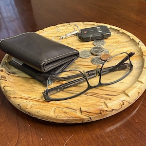 May include: A round, wooden tray with a natural, light-colored finish holds a black leather wallet, eyeglasses, car keys, and loose change. The tray's rim has a carved, wavy design. The items are arranged on a dark wooden surface.