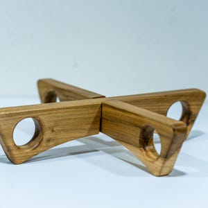 May include: A wooden plant stand with a cross-shaped design. The stand is made of light brown wood and features four triangular arms with circular cutouts. The stand is designed to hold a plant pot.