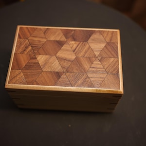 May include: A rectangular wooden box with a geometric pattern on the lid. The lid is made of small, triangular pieces of wood arranged in a repeating pattern. The box is made of light-colored wood with a darker wood lid.