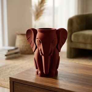 K&ouml;nnte beinhalten: Eine terrakottafarbene Vase in Elefantenform. Die Vase hat gro&szlig;e Ohren, einen R&uuml;ssel und sitzt aufrecht. Die Vase steht auf einem Holztisch in einem Wohnzimmer. Es ist ein dekorativer Gegenstand.