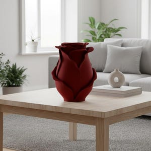 以下が含まれることがあります： 深紅のバラの形をした花瓶が、明るい木製のコーヒーテーブルの上に置かれています。花瓶は焦点であり、花びらは完全に開いています。小さなベージュの装飾品と本の山もテーブルにあります。背景には灰色のソファと植物があります。