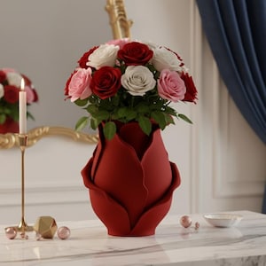 May include: A red, rose-shaped vase filled with a bouquet of red, white, and pink roses. The vase sits on a white marble table, with a gold candle holder and decorative objects nearby. A mirror and blue curtain are in the background.