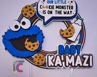 May include: A blue Cookie Monster-themed baby shower decoration. The design includes a speech bubble with the text "OUR LITTLE COOKIE MONSTER IS ON THE WAY," cookies, and the name "BABY KA'MAZI."