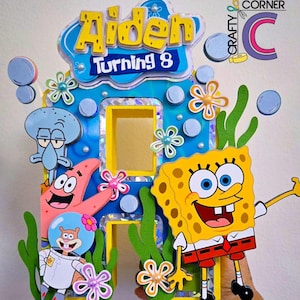 May include: A yellow and blue cardboard letter 'A' decorated with SpongeBob SquarePants characters, including SpongeBob, Patrick Star, and Squidward. The letter is decorated with blue and white bubbles, yellow flowers, and green seaweed. The letter says "Aiden Turning 8" at the top.