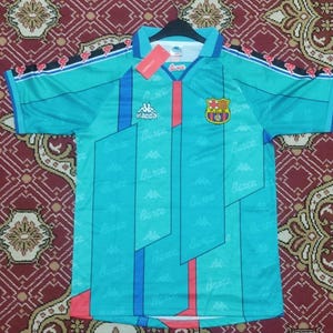May include: A turquoise soccer jersey with a red, white, and blue geometric pattern. The jersey has the Kappa logo and the Barcelona crest on the front. The sleeves have red, white, and blue stripes with the Kappa logo on the shoulders.