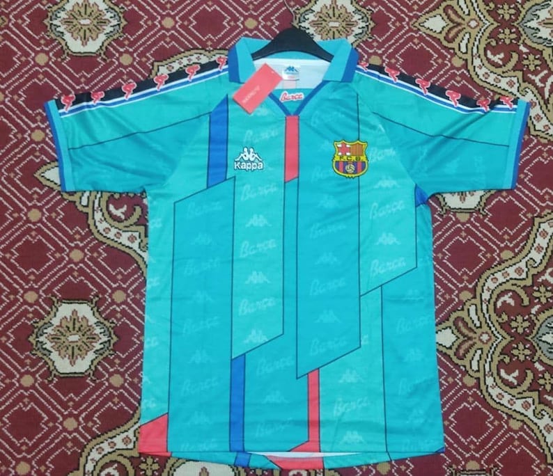 May include: A turquoise soccer jersey with a red, white, and blue geometric pattern. The jersey has the Kappa logo and the Barcelona crest on the front. The sleeves have red, white, and blue stripes with the Kappa logo on the shoulders.