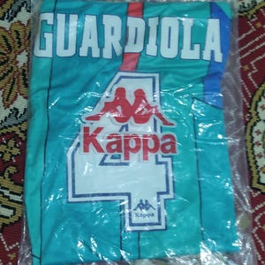 May include: A teal blue soccer jersey with white and red accents. The jersey has the name "GUARDIOLA" printed on the front in white letters with a red outline. The Kappa logo is printed on the front of the jersey in red and white.