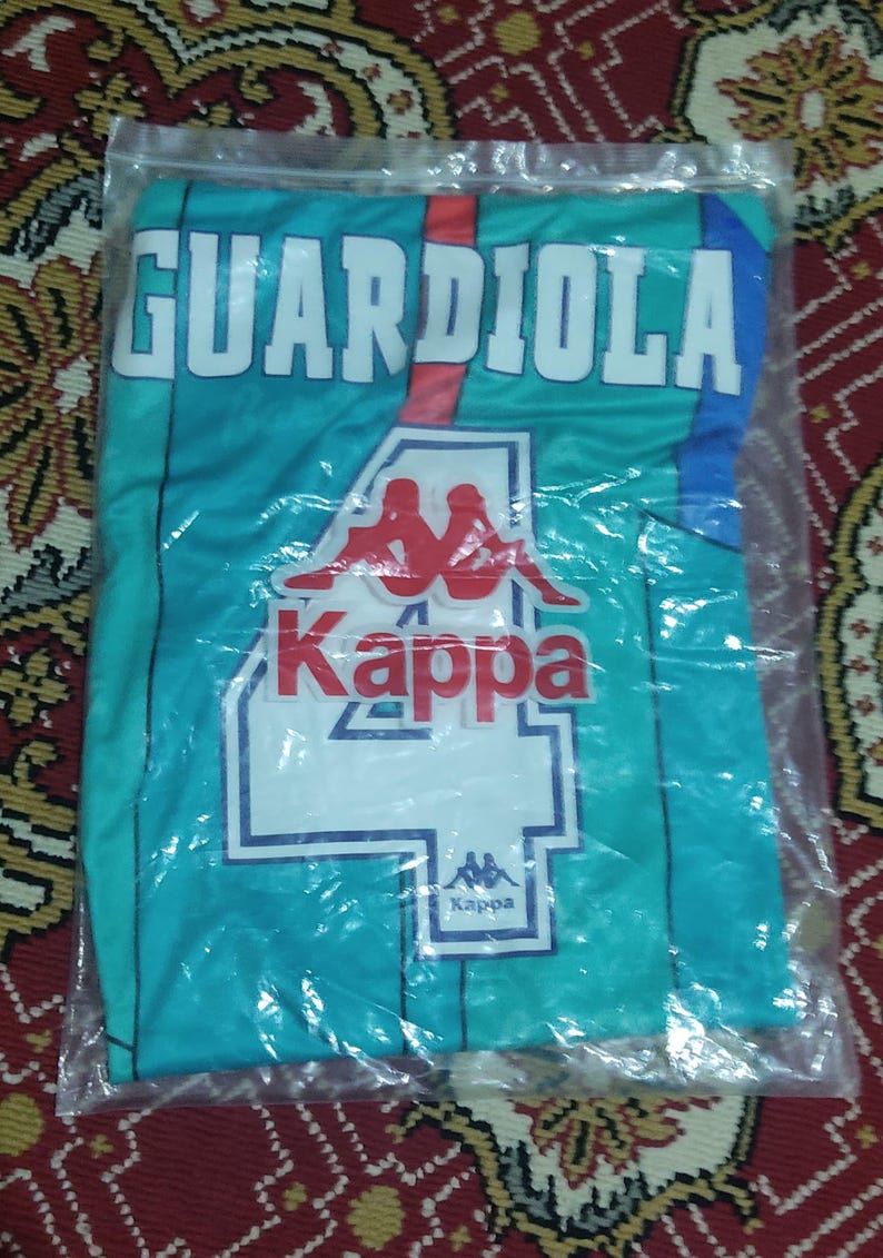 May include: A teal blue soccer jersey with white and red accents. The jersey has the name "GUARDIOLA" printed on the front in white letters with a red outline. The Kappa logo is printed on the front of the jersey in red and white.
