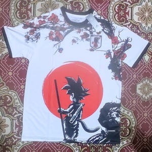 May include: White t-shirt featuring a red sun graphic with a black cartoon character holding a staff. The shirt has a floral design with red flowers and black accents. The Japanese team emblem is visible on the upper chest.