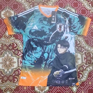 以下が含まれることがあります： アニメのキャラクターと日本の国旗が描かれた、オレンジと黒の半袖グラフィックTシャツ。Vネックで、アディダスのロゴが入っています。「HEAT.RDY」の文字が見えます。