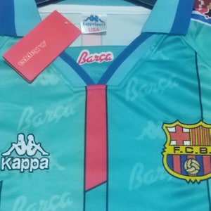 May include: A turquoise blue soccer jersey with blue and white stripes on the sleeves. The jersey has the Kappa logo on the left chest and the FC Barcelona logo on the right chest. The jersey is printed with the word "Barca" in white.