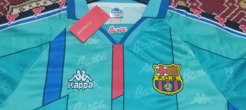 May include: A turquoise blue soccer jersey with blue and white stripes on the sleeves. The jersey has the Kappa logo on the left chest and the FC Barcelona logo on the right chest. The jersey is printed with the word "Barca" in white.
