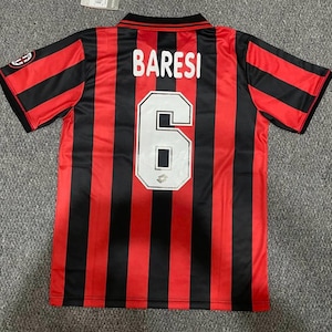 Pode incluir: Uma camisola de futebol de manga curta com riscas verticais vermelhas e pretas. As costas apresentam o nome BARESI em letras brancas sobre o número 6 numa fonte branca grande com um contorno preto. Um logótipo é visível na manga.