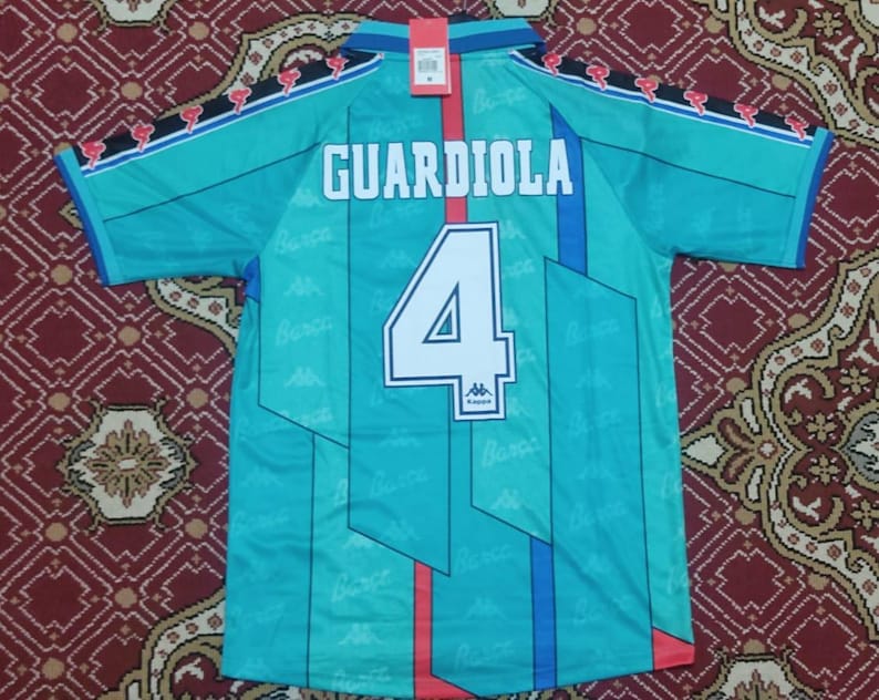 May include: A teal blue soccer jersey with a white number 4 and the name "GUARDIOLA" on the back. The jersey has a geometric pattern and red and blue accents. The jersey is made by Kappa.