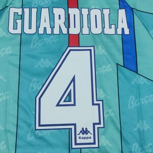 May include: A blue and white soccer jersey with the name "GUARDIOLA" and the number "4" on the back. The jersey has a Kappa logo and the word "Barca" repeated on the fabric.