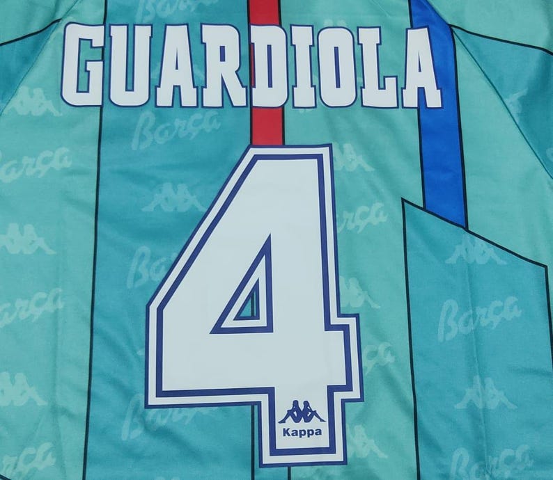 May include: A blue and white soccer jersey with the name "GUARDIOLA" and the number "4" on the back. The jersey has a Kappa logo and the word "Barca" repeated on the fabric.