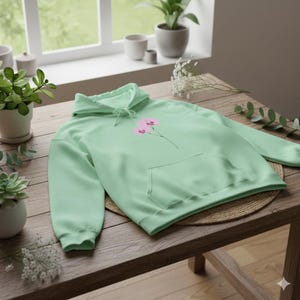 Pode incluir: Um moletom verde claro com um bolso frontal e um capuz com cordão. O moletom apresenta um design floral rosa na frente. A peça de roupa é feita de um material macio e é exibida em uma mesa de madeira.