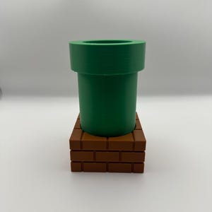 May include: A green pipe, reminiscent of a video game, sits atop a brown brick base. The pipe is cylindrical with a wide rim. The brick base is rectangular and made of individual bricks.
