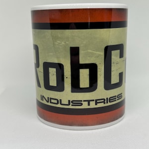 May include: White ceramic mug with a red, black, and yellow design. The mug features the text "RobCo Industries" in black.