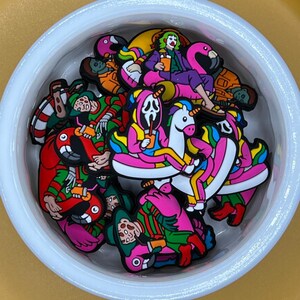 Puede incluir: Una colección de abalorios de goma de colores con varios personajes, entre ellos un payaso montando un flamenco, un fantasma montando un unicornio y un personaje de una película de terror montando un flamenco. Los abalorios están en un cuenco blanco.