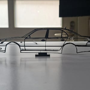 May include: A black wireframe sculpture of a saloon car silhouette, viewed from the side profile. The structure features thin lines outlining the body, windows, doors, and headlights, mounted on a small rectangular base for stability.