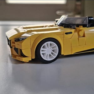 Könnte beinhalten: Ein gelbes Spielzeug-Sportauto aus Kunststoff-Bausteinen. Das Fahrzeug verfügt über ein flaches Design, eine transparente Windschutzscheibe, schwarze Innensitze und weiße Vielspeichenräder mit schwarzen Reifen. Die Frontpartie zeigt kantige Scheinwerfer und eine geformte Motorhaube.