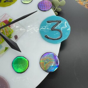 May include: A white clock face with a black hand and colorful glass numbers and accents. The number 3 is a turquoise blue circle with a black 3 inside. The clock face is surrounded by a white border.