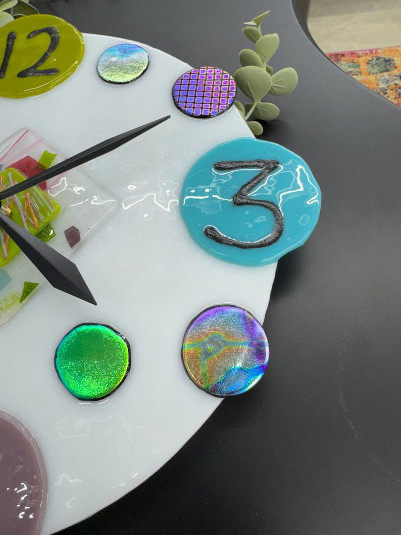 May include: A white clock face with a black hand and colorful glass numbers and accents. The number 3 is a turquoise blue circle with a black 3 inside. The clock face is surrounded by a white border.