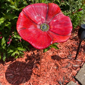 May include: A red ceramic poppy flower with a yellow center, mounted on a metal stake. The flower is sitting on a bed of red mulch.