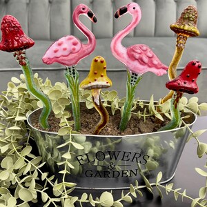 May include: A set of six glass garden stakes featuring pink flamingos and colorful mushrooms. The stakes are arranged in a metal planter with the words "Flowers & Garden" printed on the side.