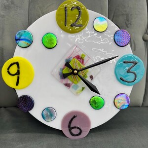 May include: A white circular clock face with colorful glass accents and black numbers. The clock face has a green, pink, and white mosaic design in the center. The clock hands are black.
