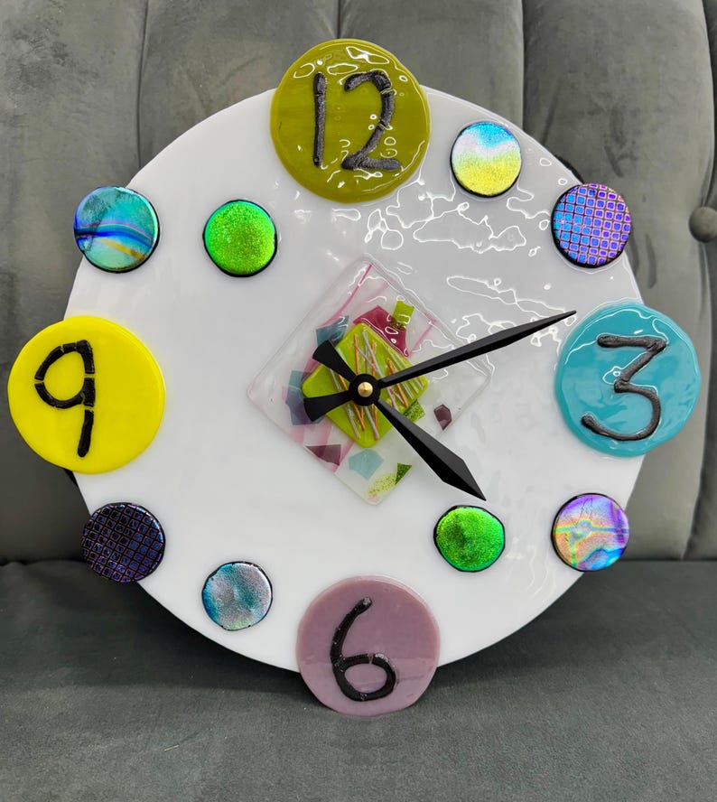 May include: A white circular clock face with colorful glass accents and black numbers. The clock face has a green, pink, and white mosaic design in the center. The clock hands are black.