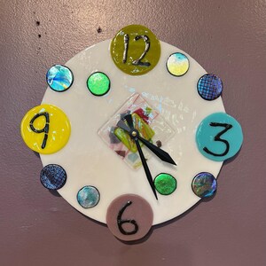 May include: A white clock face with colorful glass numbers and a mosaic design in the center. The clock hands are black and the numbers are 12, 9, 6, and 3.