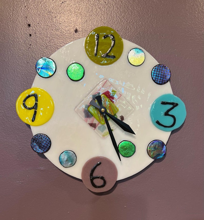 May include: A white clock face with colorful glass numbers and a mosaic design in the center. The clock hands are black and the numbers are 12, 9, 6, and 3.