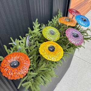 May include: A planter box filled with artificial greenery and colorful, decorative flower stakes. The flowers are in shades of red, orange, yellow, green, blue, and purple, each with a dark center. The planter is dark gray.