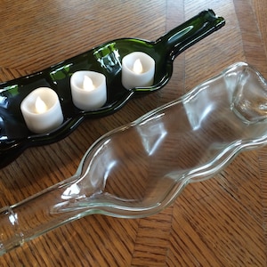 Puede incluir: Dos portavelas de vidrio hechos de botellas de vino recicladas. Uno es verde y el otro es transparente. Ambos tienen una forma ondulada y orgánica. Tres velas de té blancas se colocan en el portavelas verde.