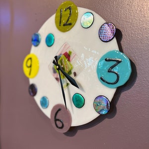May include: A white clock face with colorful glass accents and black hands. The numbers 12, 9, 3, and 6 are visible. The clock face is surrounded by a white border.